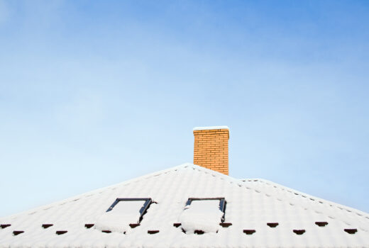 Closeup of asphalt roof covered in snow showing roof ventilation and chimney that help prevent ice dams.