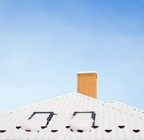 Closeup of asphalt roof covered in snow showing roof ventilation and chimney that help prevent ice dams.
